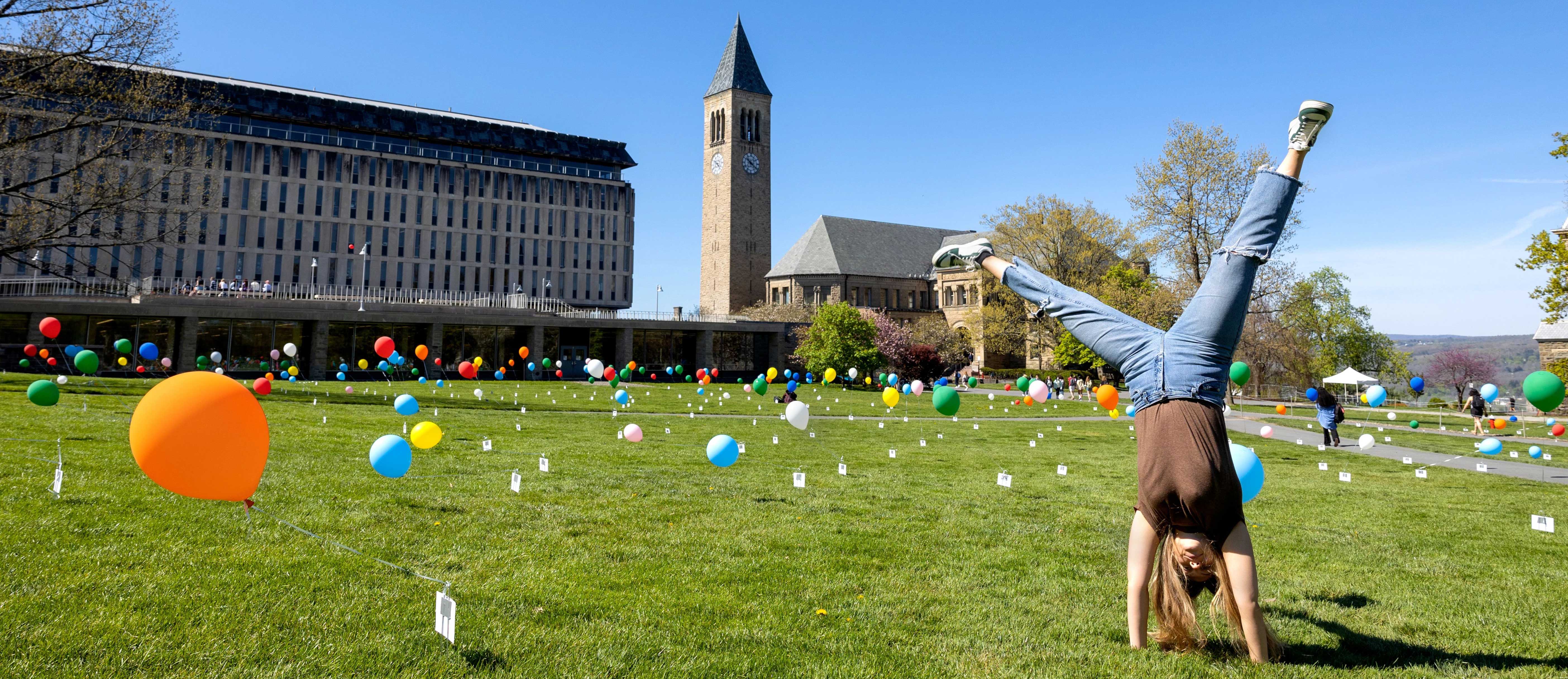 Student stands on head among colorful balloons at "Cornell Lifted" event on the Arts Quad, clocktower in background